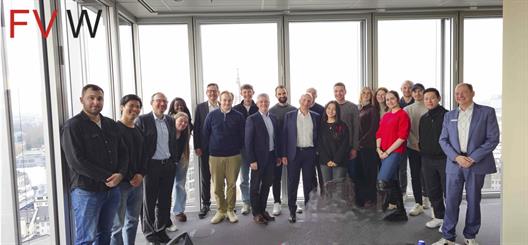 Studierende, Vortragende und Professoren stehen für ein Gruppenfoto im Halbkreis aufgereiht in einem Raum mit großem Fensterflächen, der sich in einer oberen Etage mit befindet. 